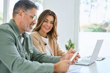 Happy middle aged mature couple using laptop counting taxes refund receipts to save money at home. Old man and woman paying bills online planning financial budget savings calculating payment at table. - obrazy, fototapety, plakaty