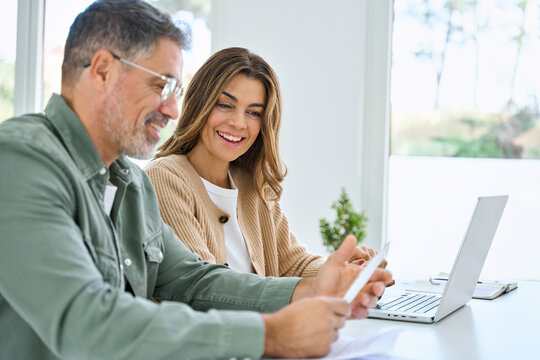 Happy middle aged mature couple using laptop counting taxes refund receipts to save money at home. Old man and woman paying bills online planning financial budget savings calculating payment at table.
