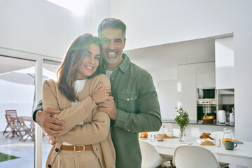 Happy romantic mature older Latin man and woman in their 50s, smiling affectionate loving middle aged couple hugging looking at camera standing together in modern kitchen at home. Portrait.