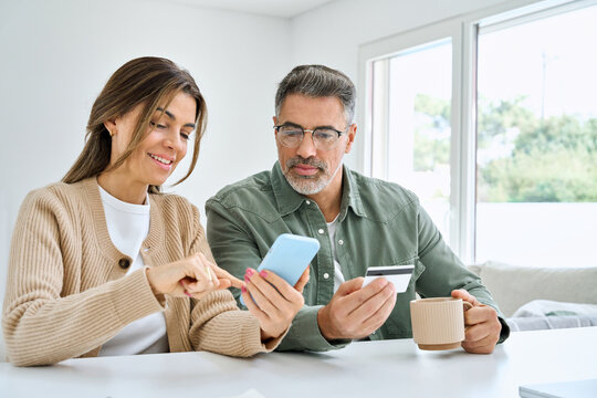 Middle aged happy couple older mature man and woman using smartphone looking at cell phone holding credit card buying online, making banking payment on ecommerce mobile shopping sitting at home table.