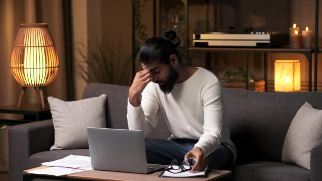 Tired Indian employee or student yawning and rubbing eyes while working or studying at home in the evening. Work fatigue, overworked entrepreneur concept.