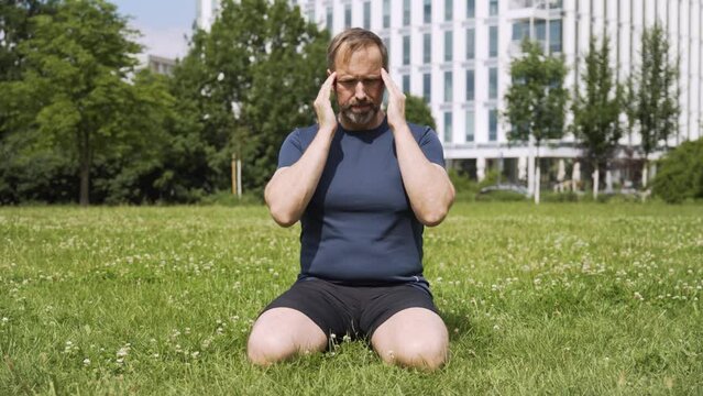 A handsome middle-aged Caucasian man rubs his head and the back of his neck as he kneels on grass in a park