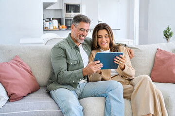 Happy middle aged couple using digital tablet relaxing on couch at home. Smiling mature man and...