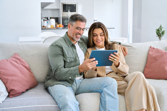 Happy middle aged couple using digital tablet relaxing on couch at home. Smiling mature man and woman holding tab computer browsing internet on pad technology device sitting on sofa in living room. - Powered by Adobe