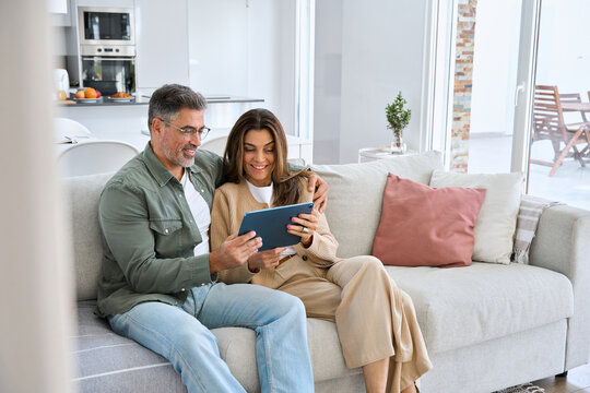 Happy middle aged couple using digital tablet relaxing on couch at home. Smiling mature man and woman holding tab browsing internet on pad device sitting on sofa in living room. Authentic candid photo