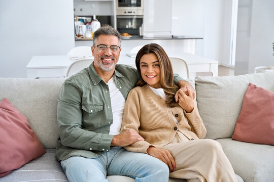 Happy smiling healthy middle aged romantic couple mature man and woman hugging sitting on sofa at home together relaxing on couch looking at camera in modern house living room. Portrait. - Powered by Adobe