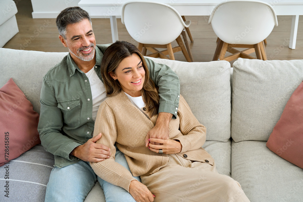 Wall mural Happy middle aged mature couple hugging looking away relaxing on couch at home. Relaxed serene older man and woman in love sitting on sofa together embracing in modern house living room. - Wall murals