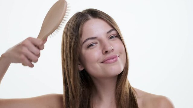 Happy caucasian lady brushing, combing hair using wooden brush standing over white background. Hair beauty, care, health and cosmetics, treatment concept.