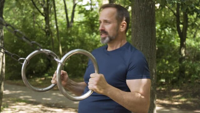 A handsome middle-aged Caucasian man does bodyweight rows with rings in a park on a sunny day