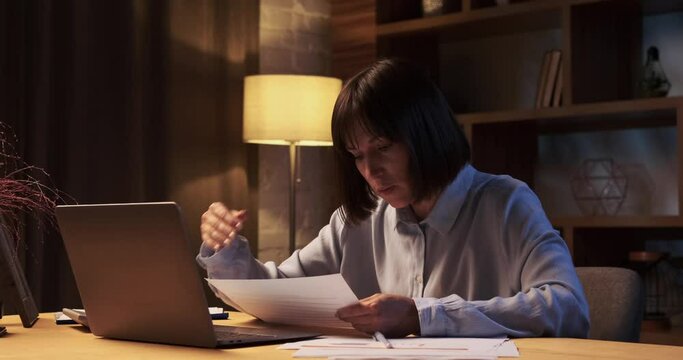 Dedicated Caucasian woman, businesswoman or freelancer working overtime on laptop computer with documents. Businesswoman burning the midnight to working hard.