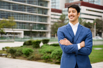 A young Hispanic businessman in a smart blue suit crosses his arms, looking off into the distance with an air of contemplation against a cityscape.