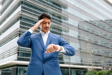 A Hispanic businessman in a blue suit looks concerned while checking the time on his watch outside a corporate building, representing the fast-paced nature of business, feels leak of time and deadline