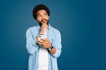 Pensive man appears intrigued as he holds his smartphone, chin resting on his hand, conveying a...