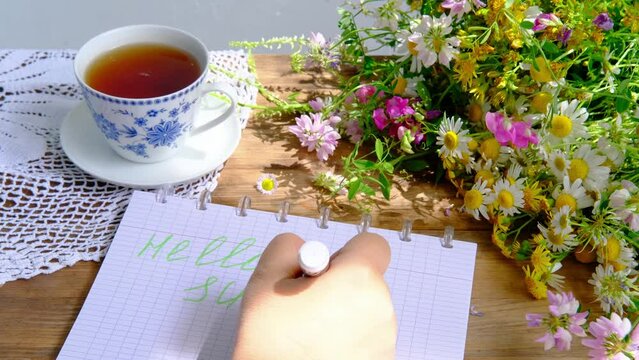female hands write hello summer, a cup of tea, a beautiful summer bouquet of wildflowers, a handmade lacy white doily on the table, the concept of summer time, a tea party in the garden