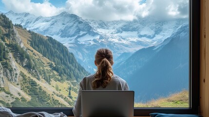 back view of a woman freelancer working on her laptop. In front of her is a large window with a green and calming forest view in a mountain.