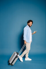 Smiling handsome Brazilian man with afro hair using a smartphone, walking with a travel suitcase. This image captures the essence of contemporary travel combined with digital communication.