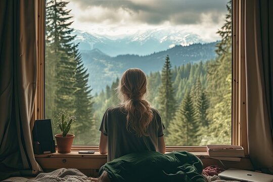 back view of a woman freelancer working on her laptop. In front of her is a large window with a green and calming forest view in a mountain.