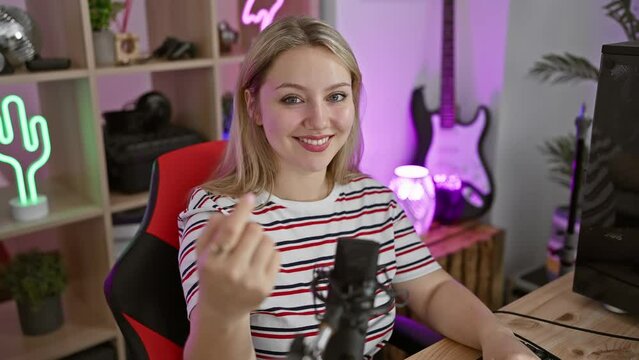 Cheerful young blonde streamer woman beckoning with her hand, inviting you to join her gaming room, happy smile lighting up the dark
