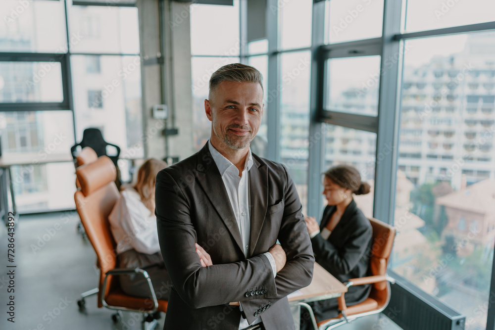 Wall mural smiling male entrepreneur with crossed hands standing in office on colleagues background