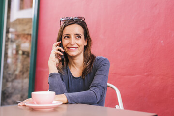 Smiling 40 year old woman using her smartphone sitting on terrace drinking coffee