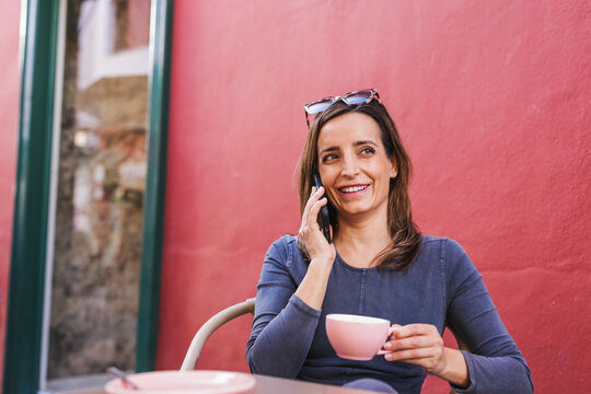 Smiling 40 Year Old Woman Using Her Smartphone Sitting On Terrace Drinking Coffee