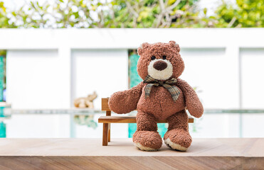 Brown teddy bear sitting on wooden bench over blurred swimming pool background, outdoor day light, valentine concept background idea