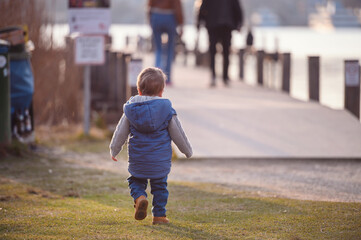 A toddler in a blue vest wanders along a lakeside path with evening light casting a warm glow