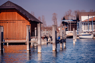 Wooden boat houses and mooring poles line a peaceful lake under a clear blue sky