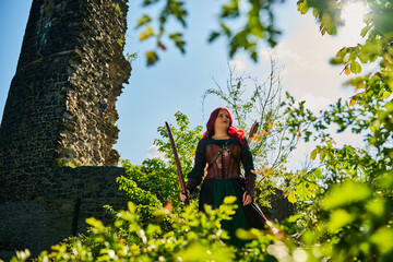 Mittelalterliche Frau mit roten Haaren als Kriegerin mit Kriegsbemalung mit Pfeil und Bogen im Sommer vor einer alten Burgruine, mit Korsett aus Leder und Umhang 