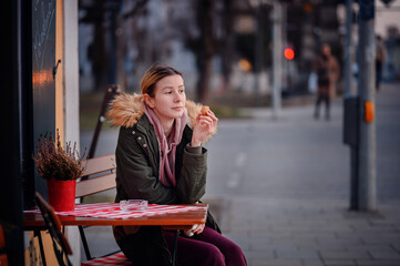 Fototapeta premium A young woman enjoys a snack at a café table on the sidewalk, with a city evening ambiance