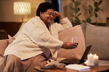 Fototapeta premium Side view portrait of senior Black woman clinking wine glass to screen during video chat with friend or long distance partner copy space