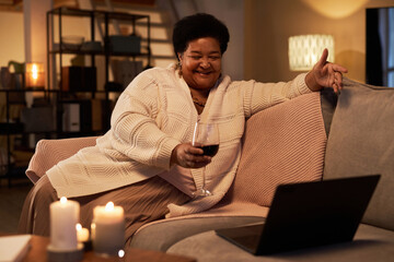 Portrait of mature Black woman smiling while enjoying conversation via video chat with glass of...