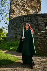 Mittelalterliche Frau mit roten Haaren als Kriegerin mit Kriegsbemalung mit Schwert im Sommer vor einer alten Burgruine, mit Korsett aus Leder und Umhang 