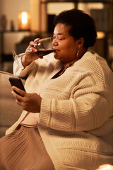 Vertical side view portrait of Black senior woman using smartphone and drinking wine during evening...