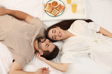 Happy couple lying on bed near white tray with breakfast, above view