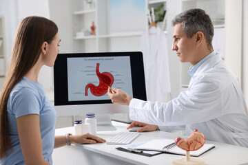 Gastroenterologist showing screen with illustration of human stomach model to patient at table in...