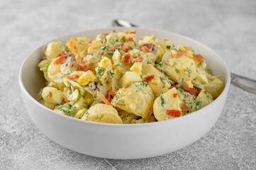 Salad of potatoes, eggs, bacon and fresh herbs with mayonnaise and yogurt dressing in a bowl on a gray concrete background.