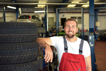Portrait of successful smiling car mechanic in a workshop on a stack of tires at his workplace - obrazy, fototapety, plakaty