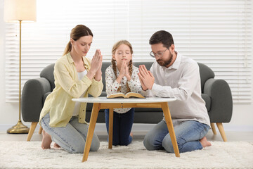 Girl and her godparents praying over Bible together at table indoors