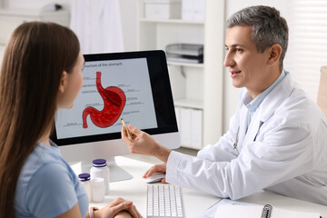 Gastroenterologist showing screen with illustration of human stomach model to patient at table in...