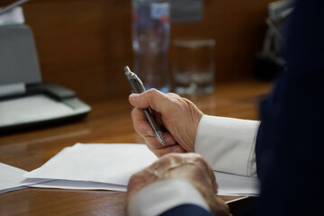 An elderly man in a business suit with cuffs, sitting at a desk with pen and documents. Concept of writing a will, drafting a contract or filling out forms. Photo. No face. Selective focus - obrazy, fototapety, plakaty