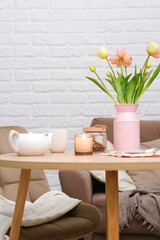 Home spring interior in living room. Pink vase with tulip,hot teapot, burning candle, cup of tea on round wooden tabl, beige chair basket in cosy living room on sofa background.