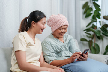 Daughter shows cancer-fighting mother something amusing on phone, joy in care. Young woman shares a cheerful moment on a smartphone with her elderly mother in a cozy room.