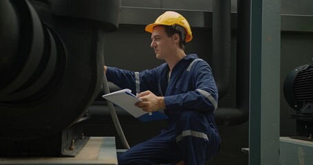 Industrial worker in blue overalls and a yellow hard hat attentively examines machinery with...