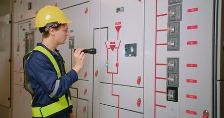 Technician in safety vest and yellow helmet using a flashlight to check electrical cabinets in a...