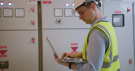 An engineer in a high-visibility vest and hard hat inspects systems on his laptop at an industrial control panel