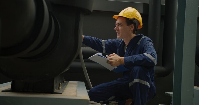 Industrial worker in blue overalls and a yellow hard hat attentively examines machinery with clipboard in hand.