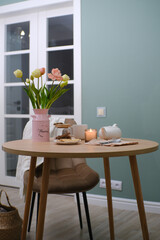 Home spring interior in living room. Pink vase with tulips and branches,hot teapot, burning candle, cup of tea on round wooden table in cosy living room.