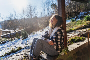 Young smiling woman in a warm blanket on the terrace of a country house enjoying the sun in early spring - 728696089