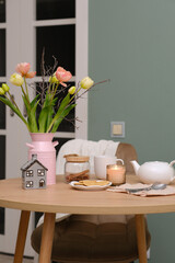 Home spring interior in living room. Pink vase with tulips and branches,hot teapot, burning candle, cup of tea on round wooden table in cosy living room.